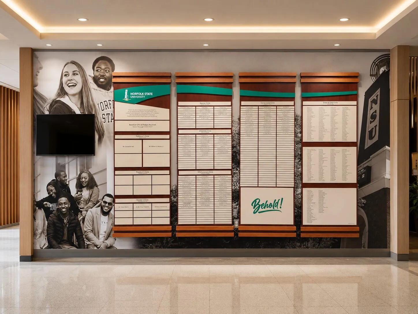 A B&D tiered donor recognition wall with rows of engraved name plaques and a bold 'Behold!' graphic, set in a university lobby beside a student photo mural.