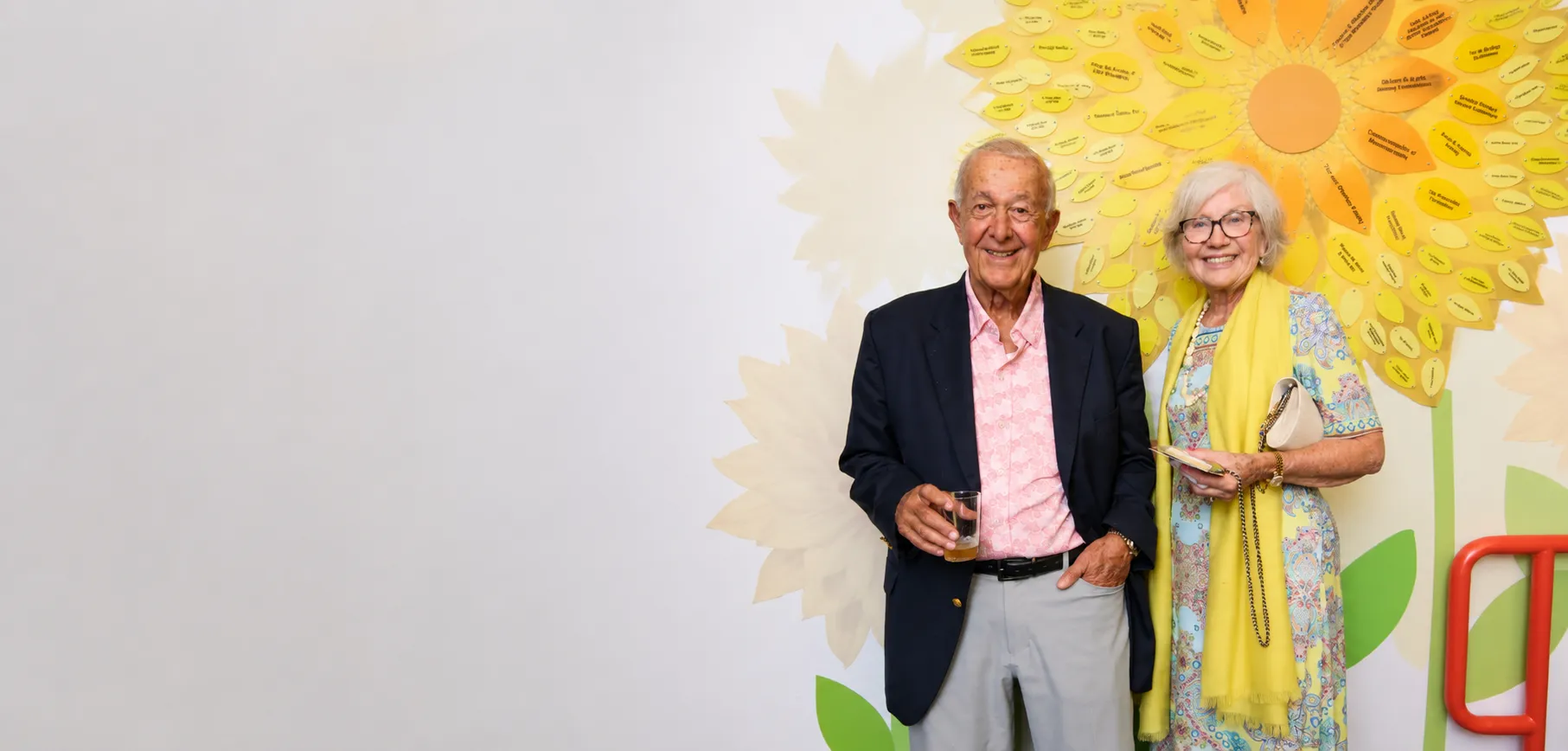 A smiling older couple standing together beside a large sunflower, representing the donors and supporters B&D has served for three decades.