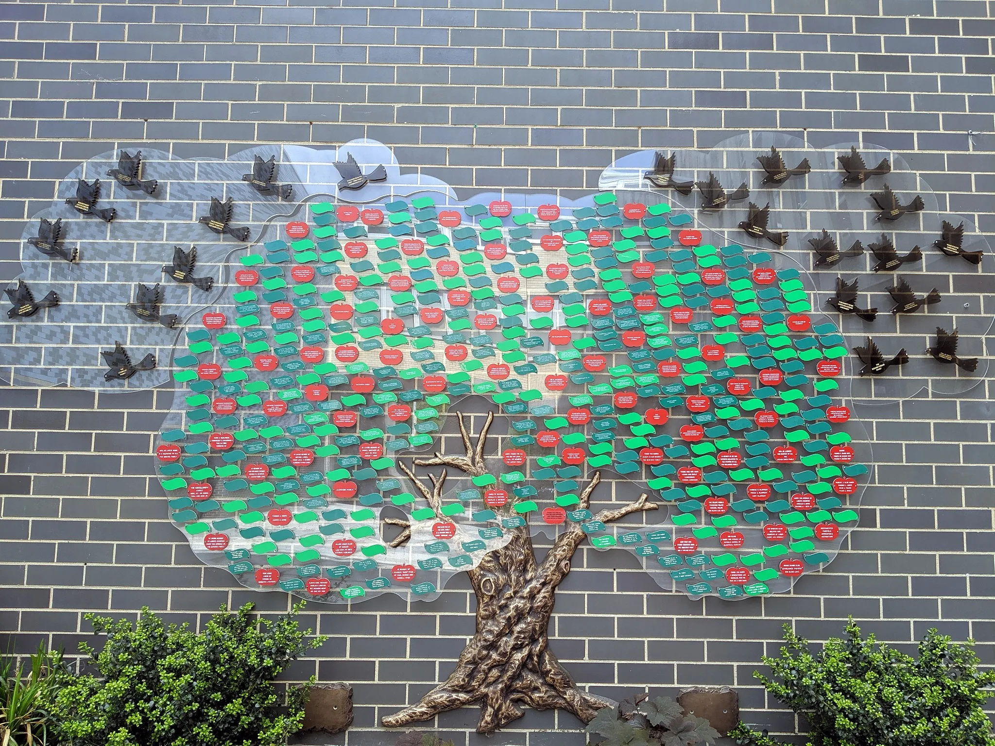 An outdoor donor tree on a brick wall, with green and red leaves and a flock of doves.