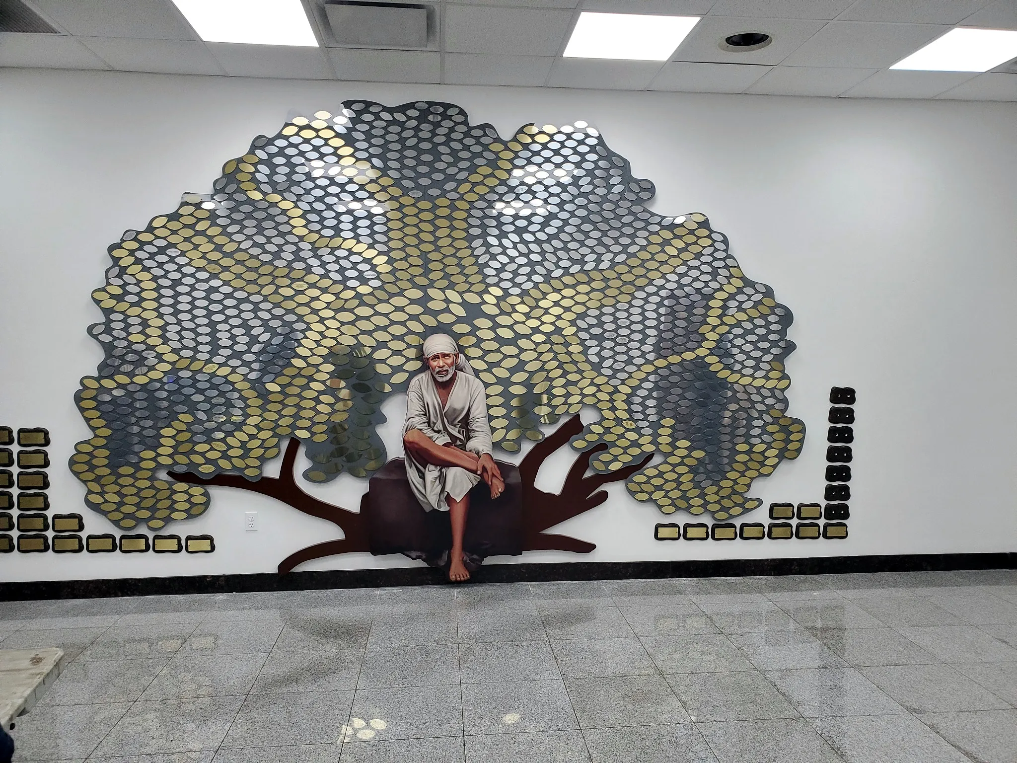 A Sri Sai donor tree with gold and silver leaves and a hand-painted figure seated at the base.