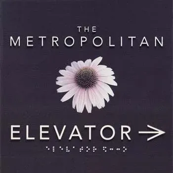 A B&D interior wayfinding sign for The Metropolitan elevator, with dimensional lettering, a directional arrow, and Braille.