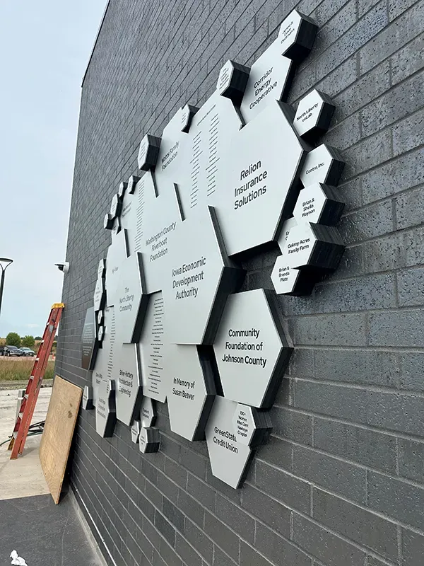 A modern B&D donor wall of layered white hexagon name plates in varied depths, mounted on a gray brick exterior wall for a community-foundation campaign.