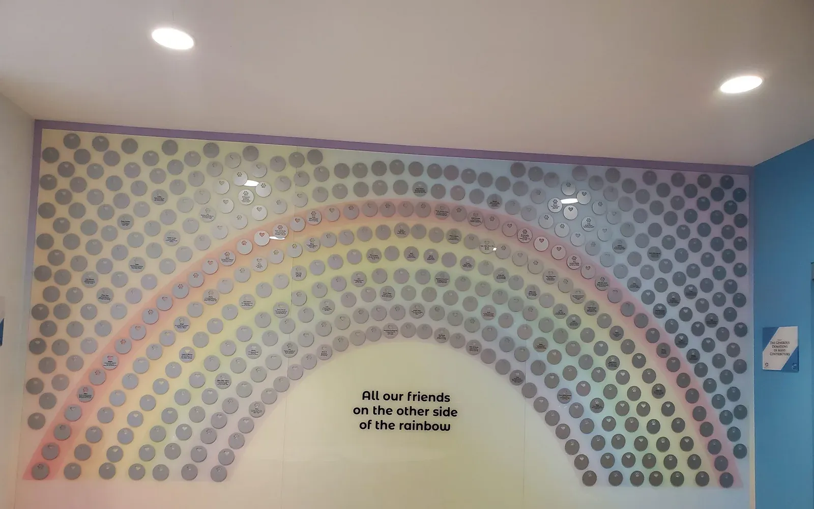 A B&D memorial display wall reading “All our friends on the other side of the rainbow,” with hundreds of engraved circular plates arranged in a rainbow arc across a pastel wall.