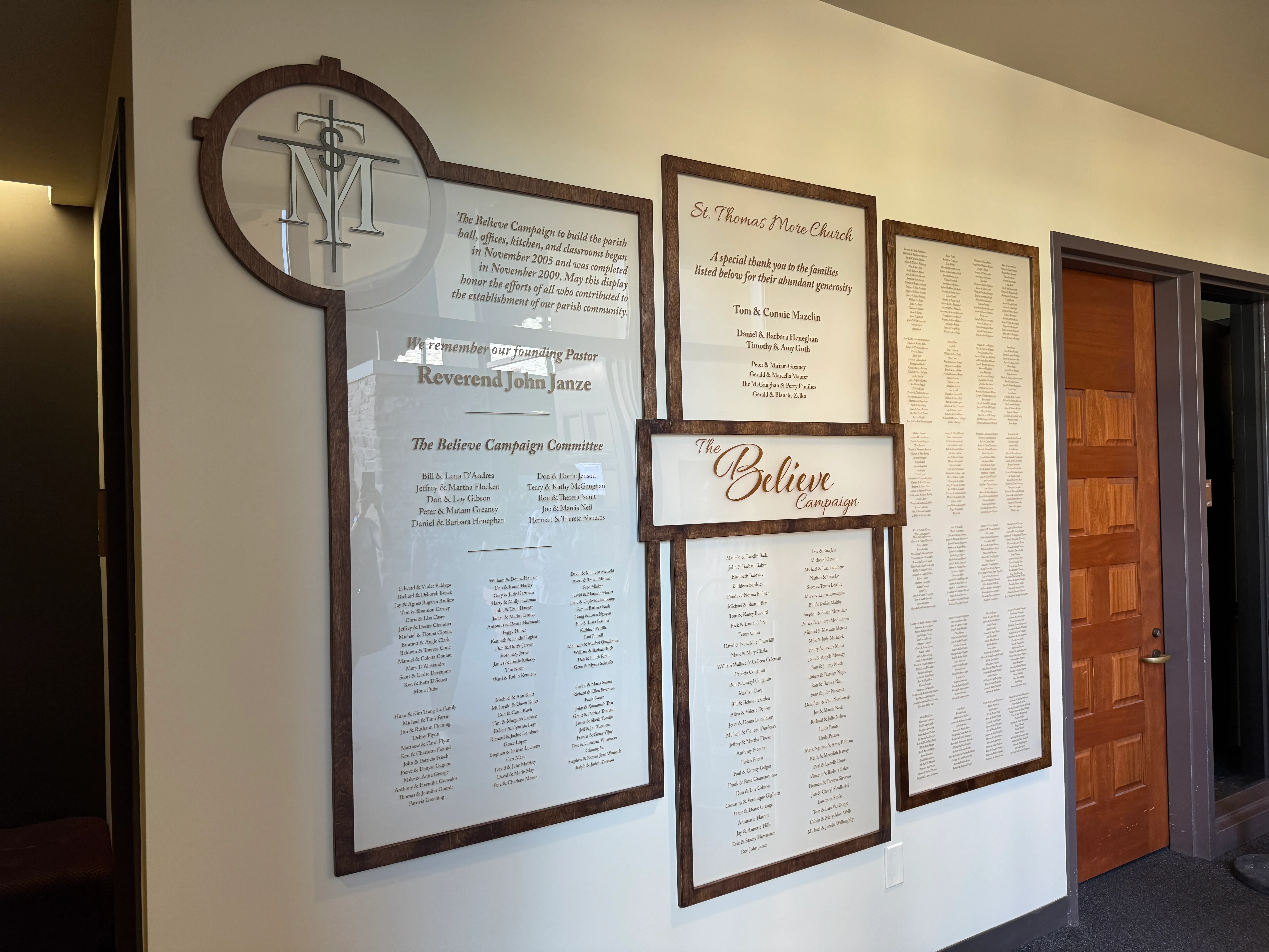 The St. Thomas More Believe Campaign donor wall in the parish hallway — wood-framed engraved name panels beside the founding-pastor tribute and the central “Believe” sign.